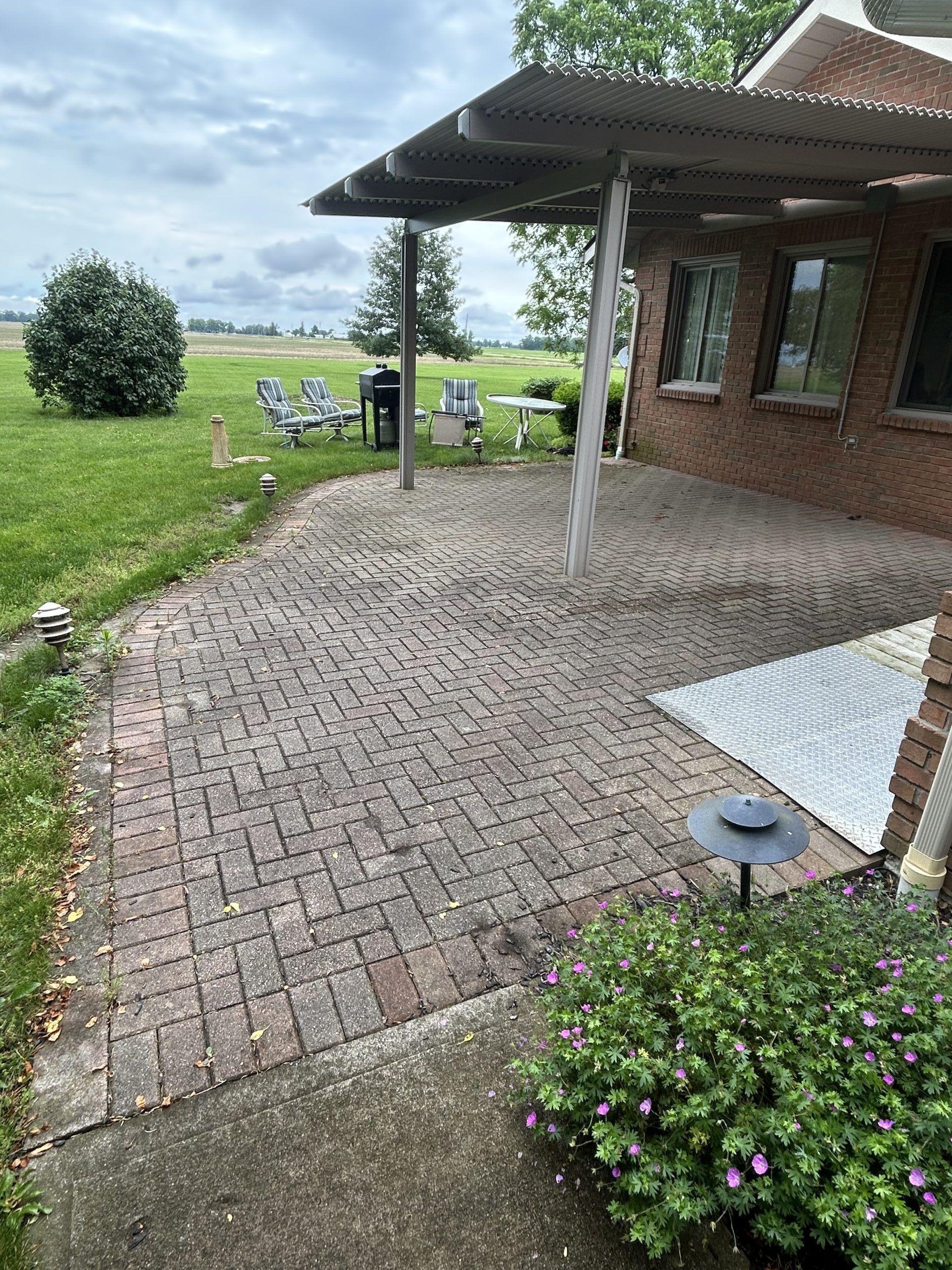Brick patio next to a brick house, with a metal awning. Green grass and a cloudy sky in the background.