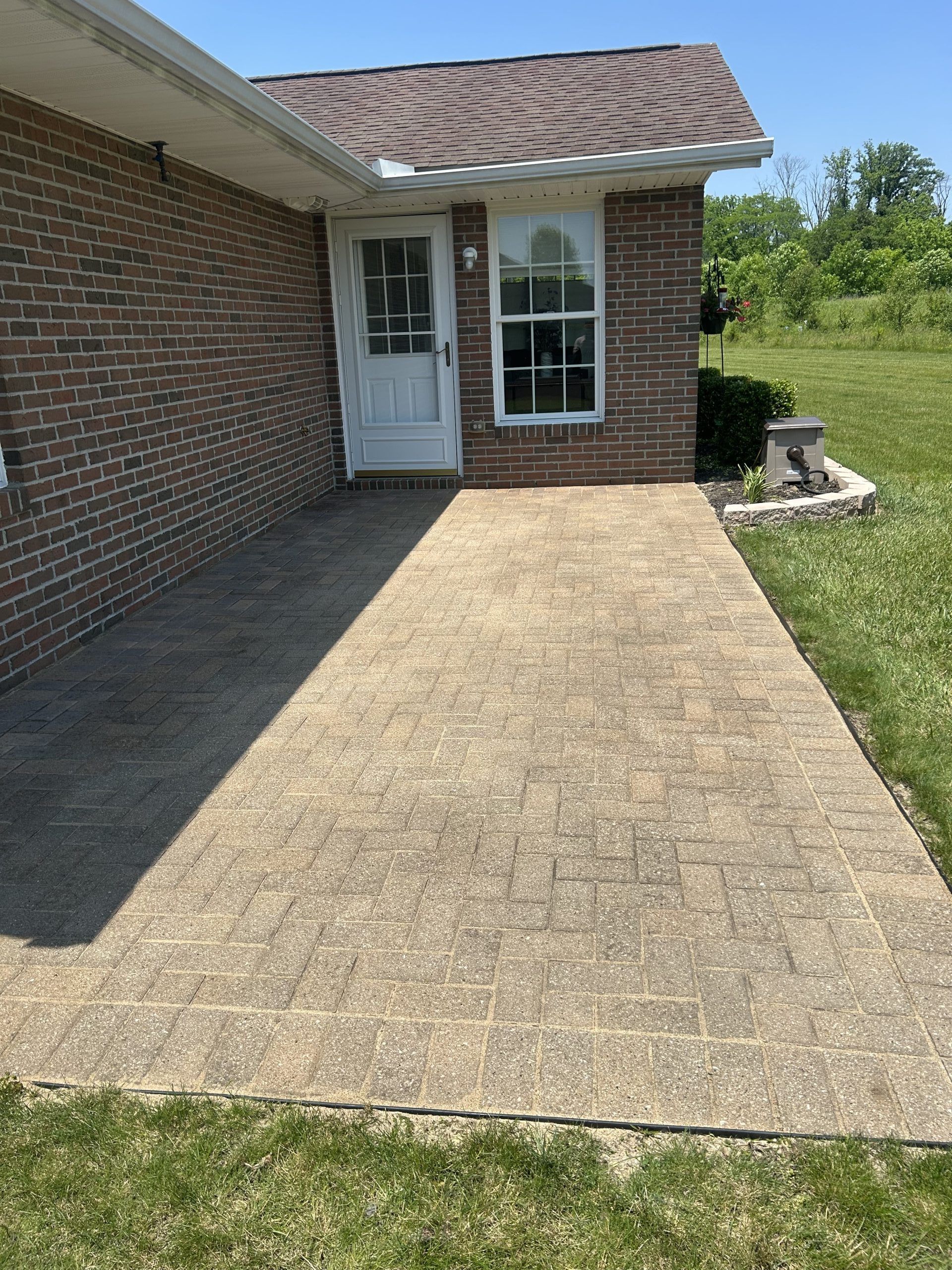 Brick-patterned concrete patio leads to a white door and window of a brick house, with green grass surrounding it.