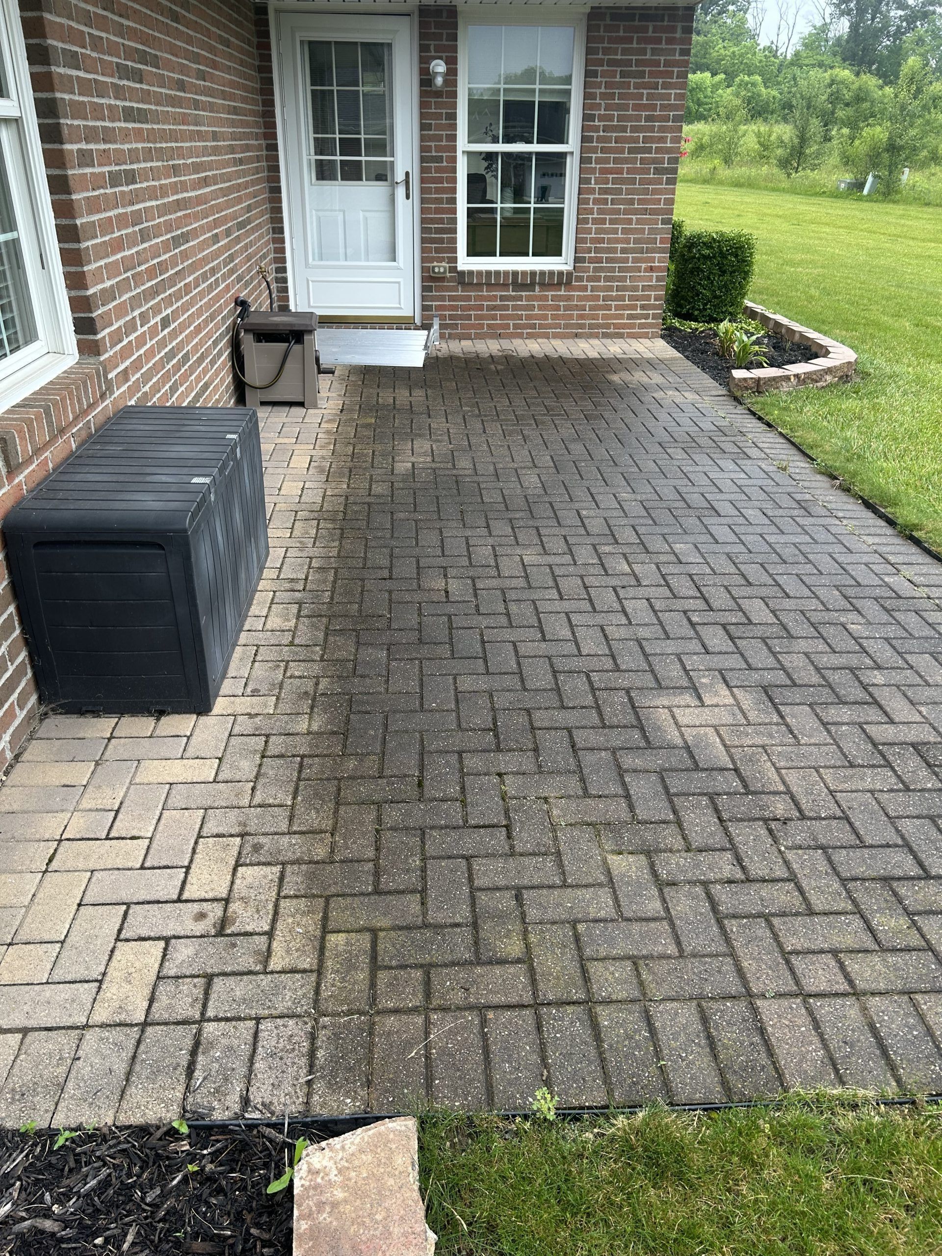 Brick patio, half cleaned, in front of a brick house. Black storage box on the left, door and window above.
