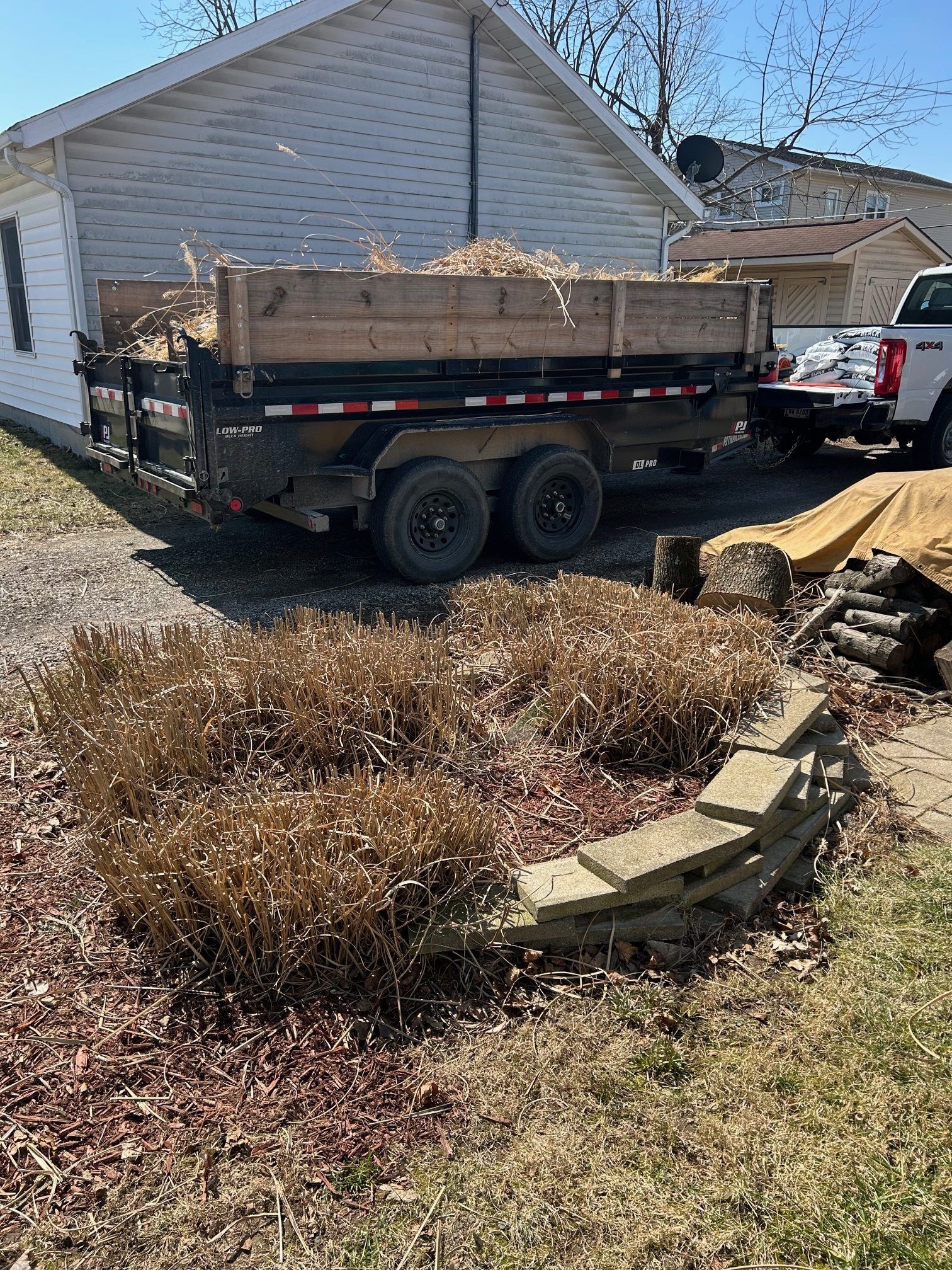 A trailer loaded with yard waste sits near a house. In the foreground are dead bushes and stacked stones.