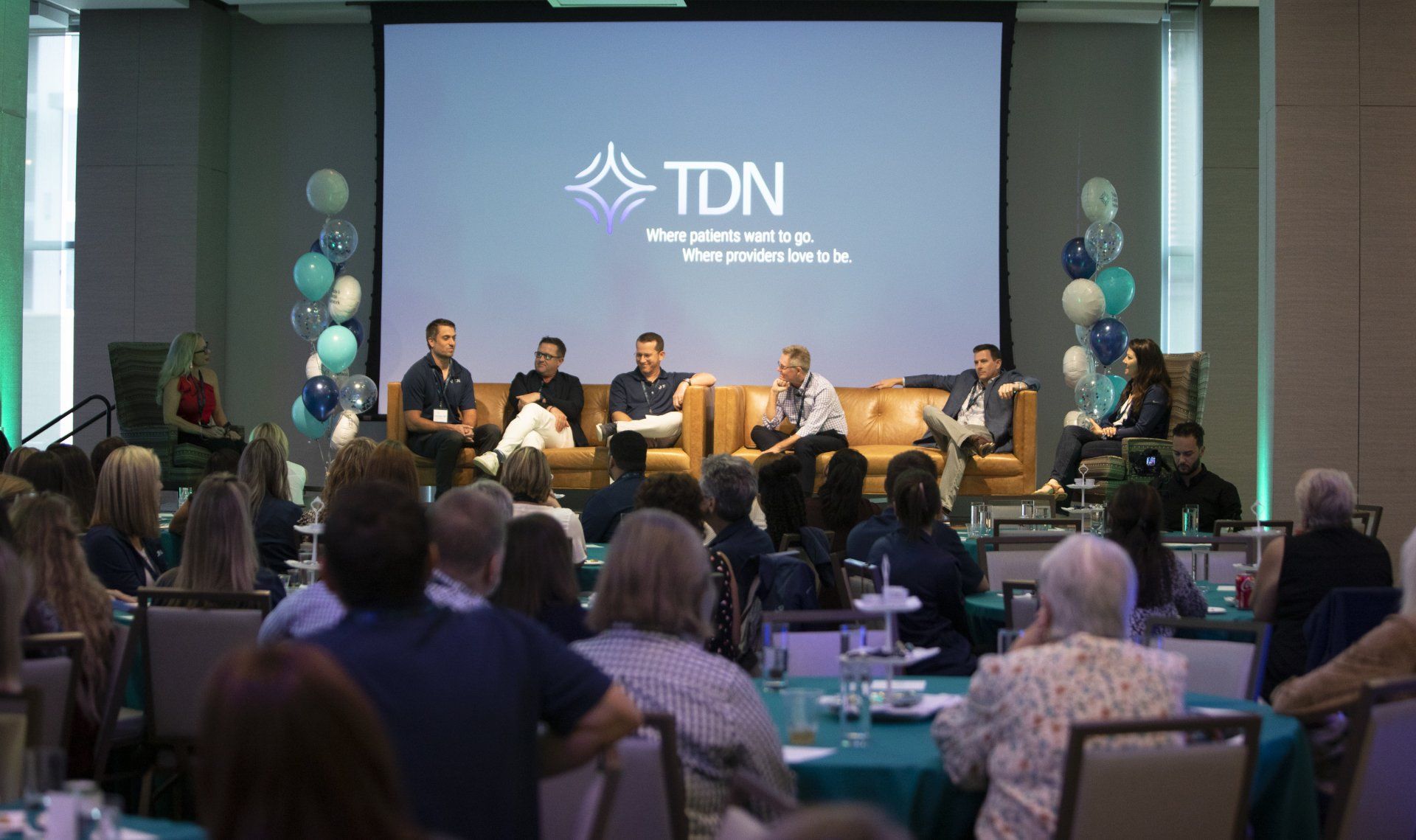 A group of people are sitting at tables in front of a large screen.