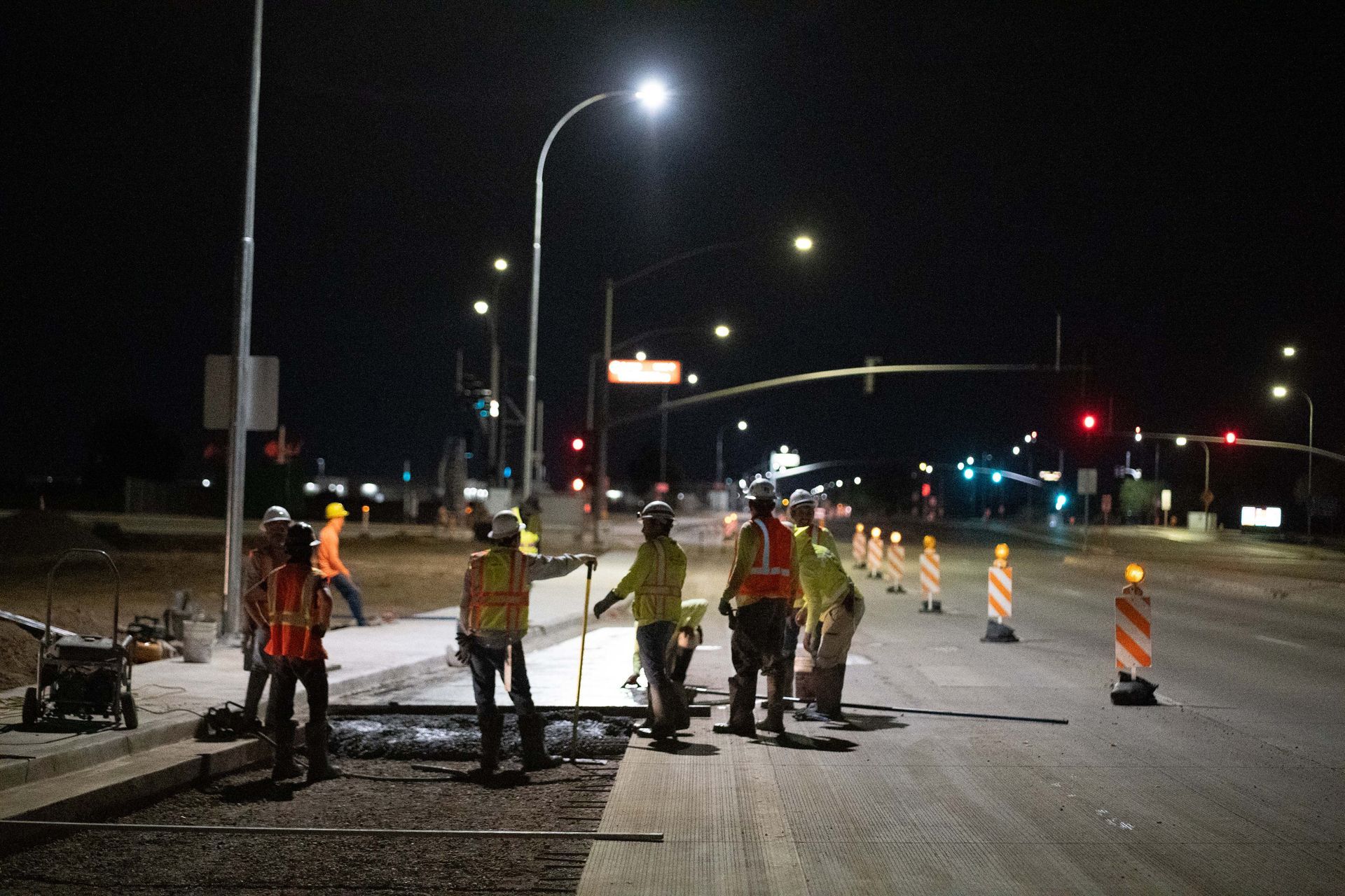 Construction workers in safety vests and helmets working on a road at night, lit by streetlights.
