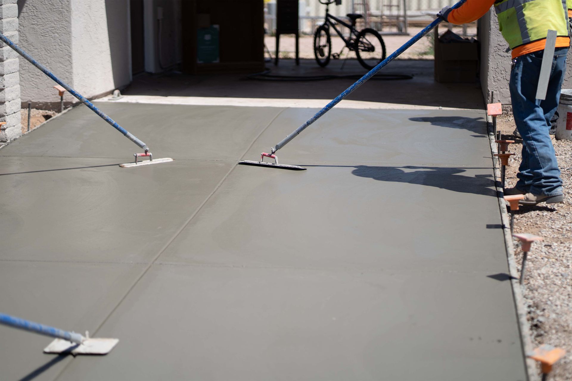 Workers smoothing wet concrete driveway with long-handled floats; bicycle in background.