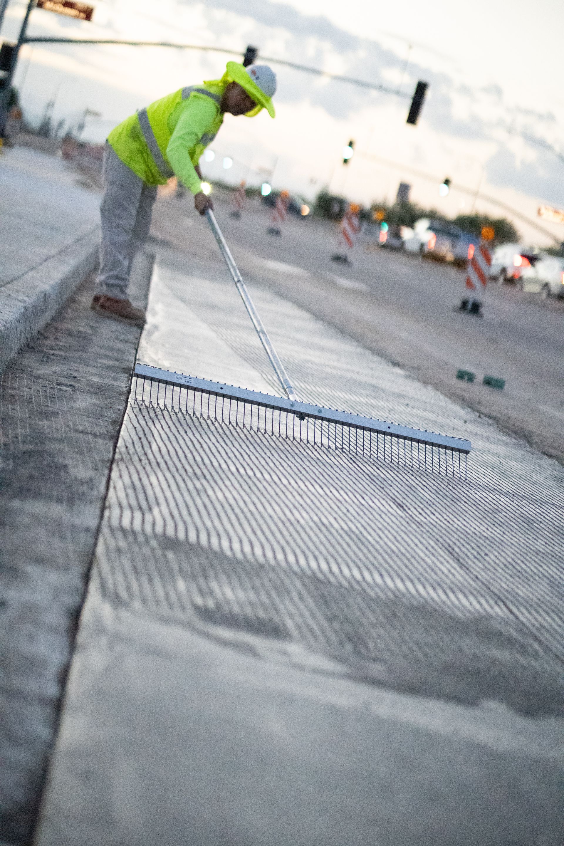 Construction worker uses a rake on freshly laid concrete on a road, wearing a neon vest and hard hat.