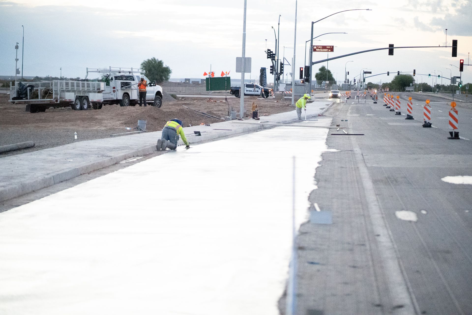 Road construction: workers applying white sealant to a roadway. Utility truck, orange cones, and traffic lights are visible.