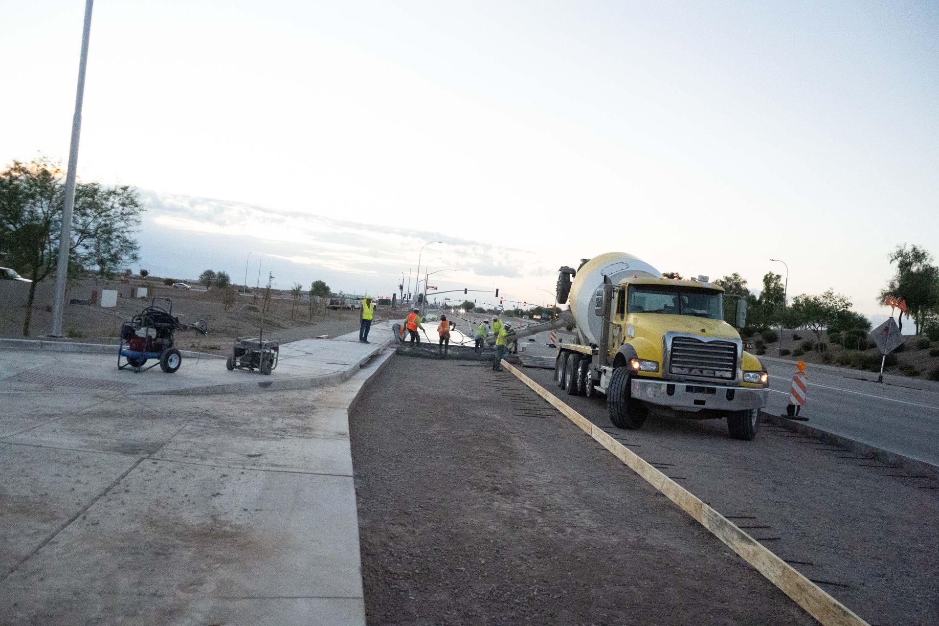 Construction site: cement truck pouring concrete on a road, workers in vests, sidewalk and road visible.