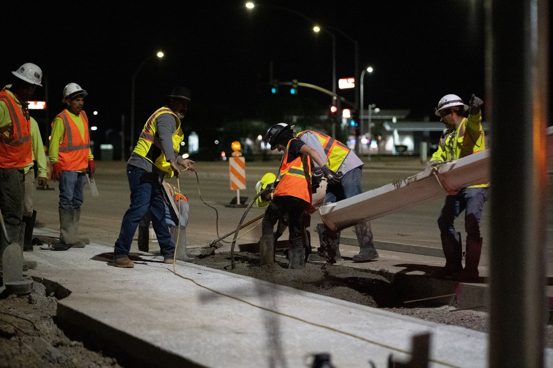 Construction workers pouring concrete at night on a street. Workers wear vests and helmets.