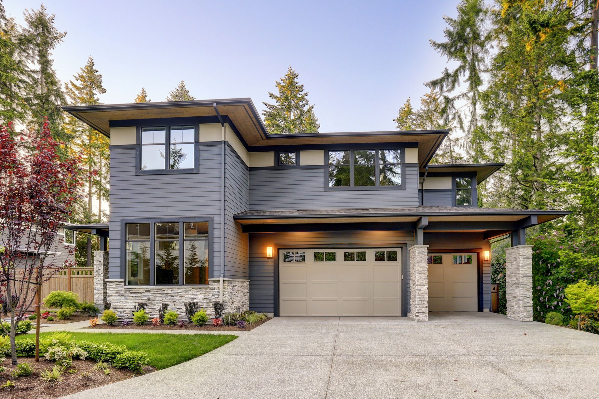 Modern two-story gray house with a two-car garage. Stone accents, brown trim, and a gravel driveway in a wooded setting.