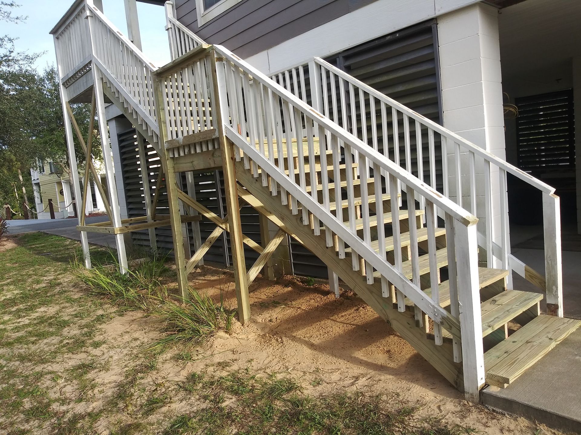 A set of outdoor wooden stairs with white railings leading up to the side of a building, built on a sandy, grassy slope.