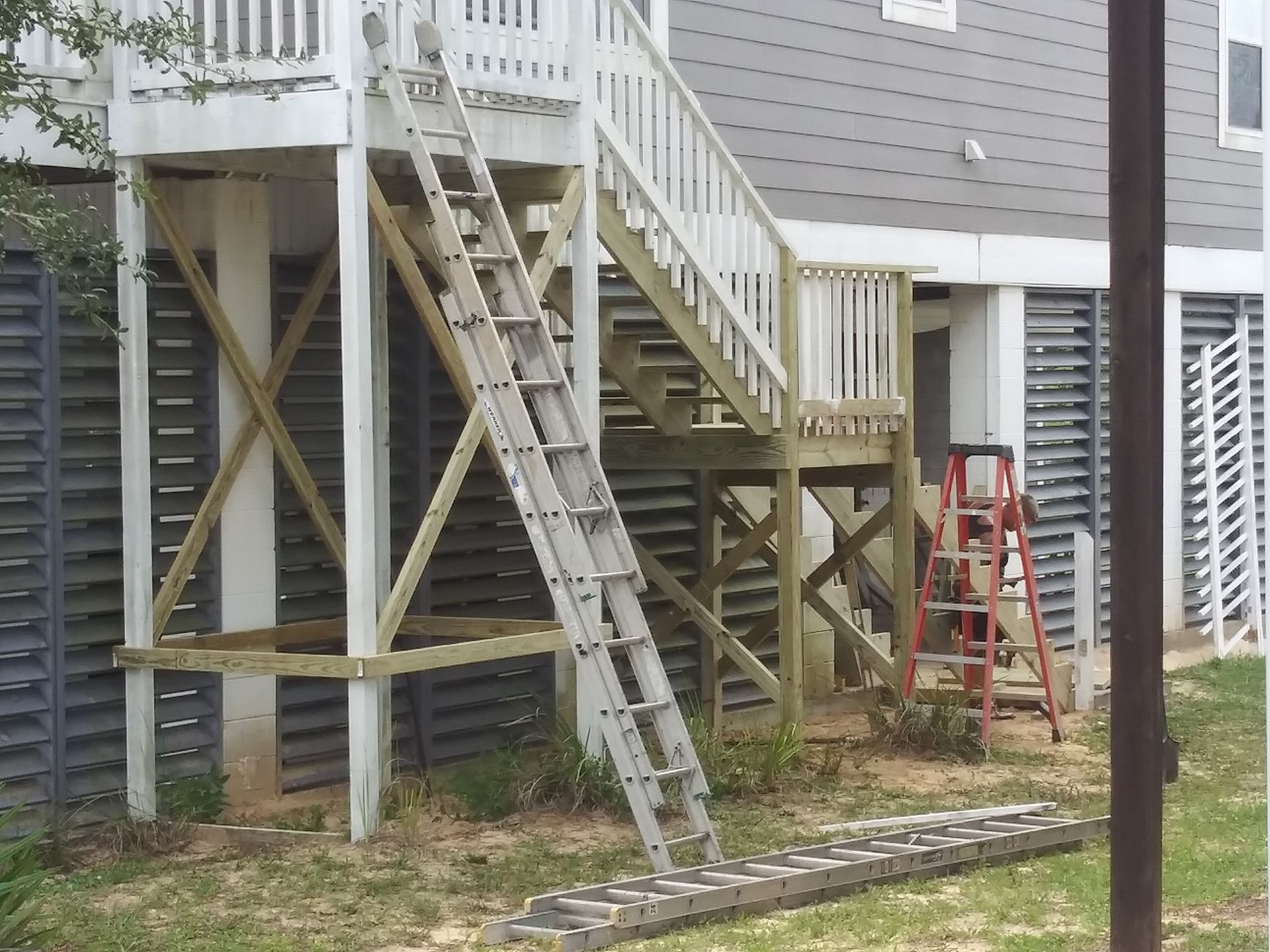 An extension ladder leans against an elevated wooden deck structure, with a red stepladder and another ladder on the ground.