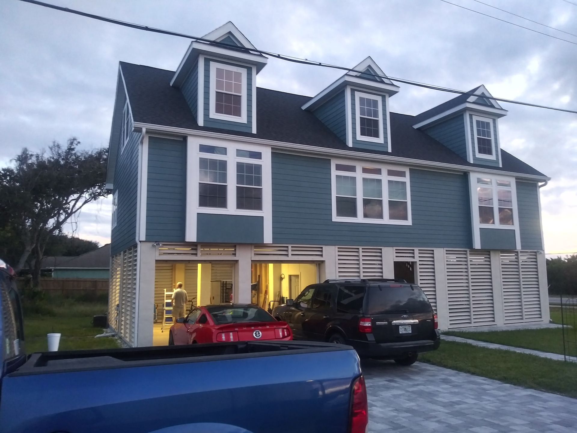 A blue two-story house with dormer windows, raised on a white foundation with a car parked in the ground-level garage.