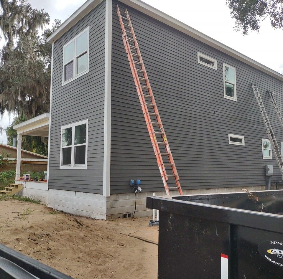A two-story house with grey siding and white trim under renovation, featuring two ladders leaning against the side.