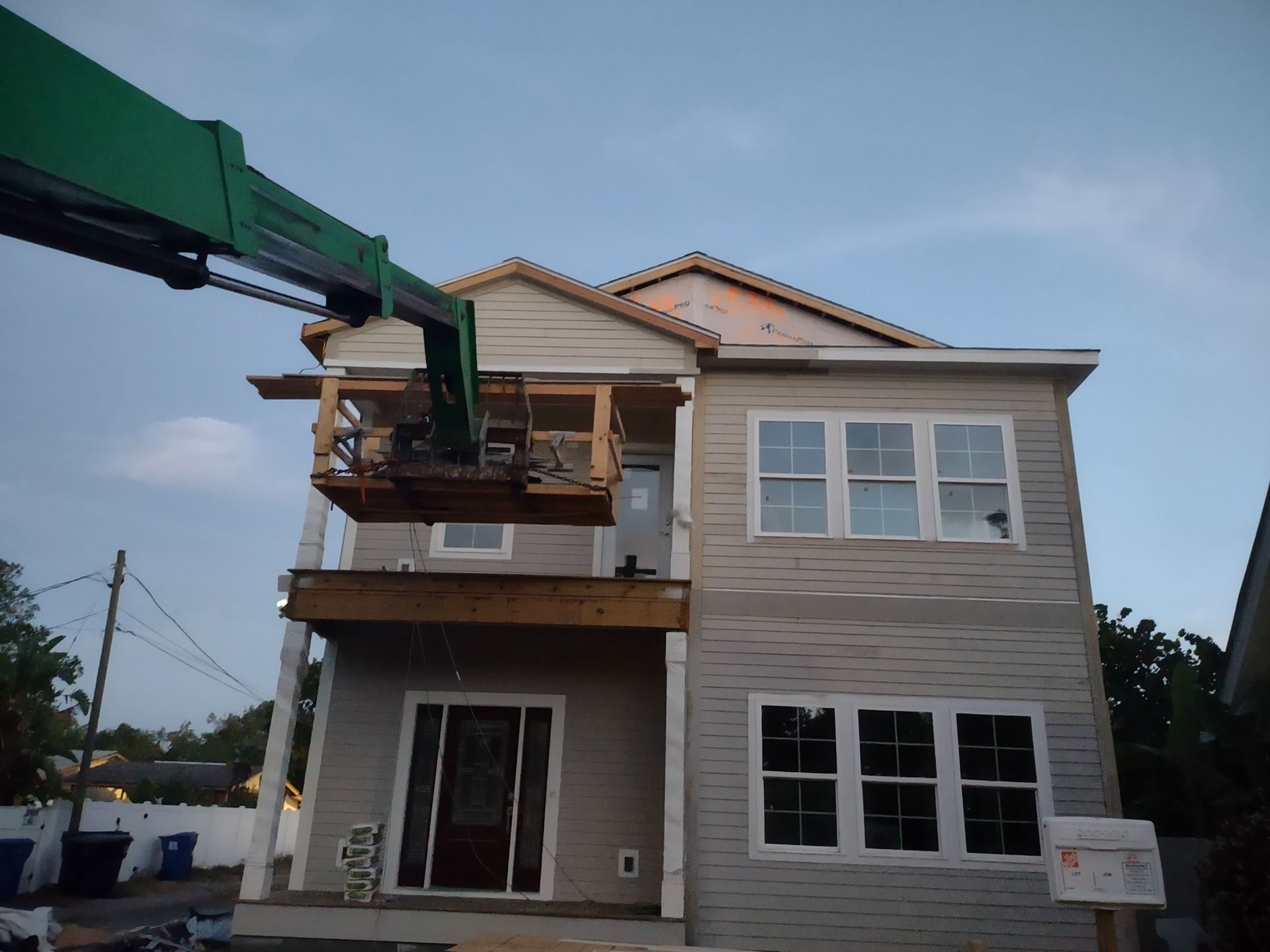A boom lift platform reaches the upper balcony of a two-story gray house under construction.