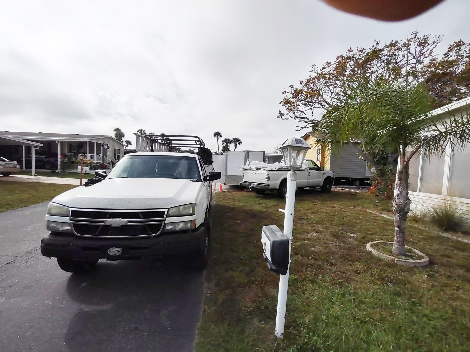A white pickup truck parked in a driveway next to a grass lawn with another truck and trailer in the background.