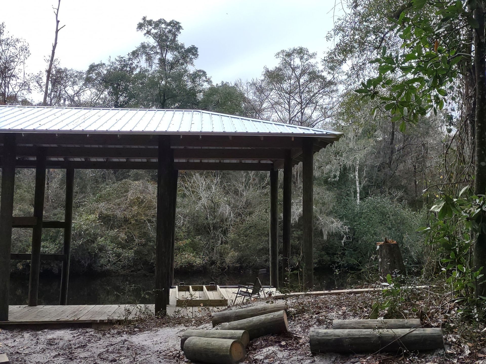 A metal-roofed pavilion structure overlooking a dark river, surrounded by trees with cut logs lying in the foreground.