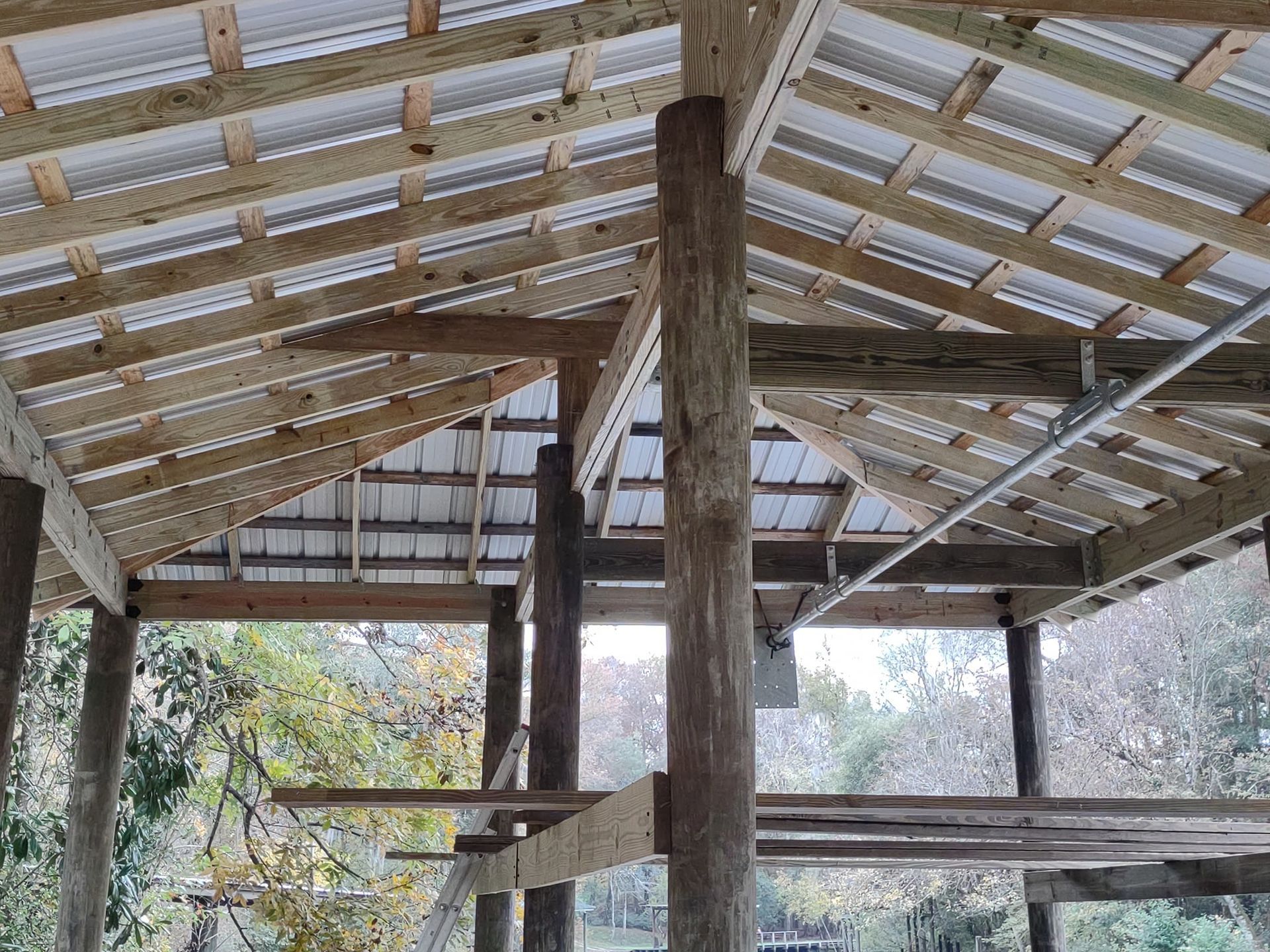 A low-angle view of a wooden post-and-beam structure with a metal roof under construction, showing rafters and support poles.