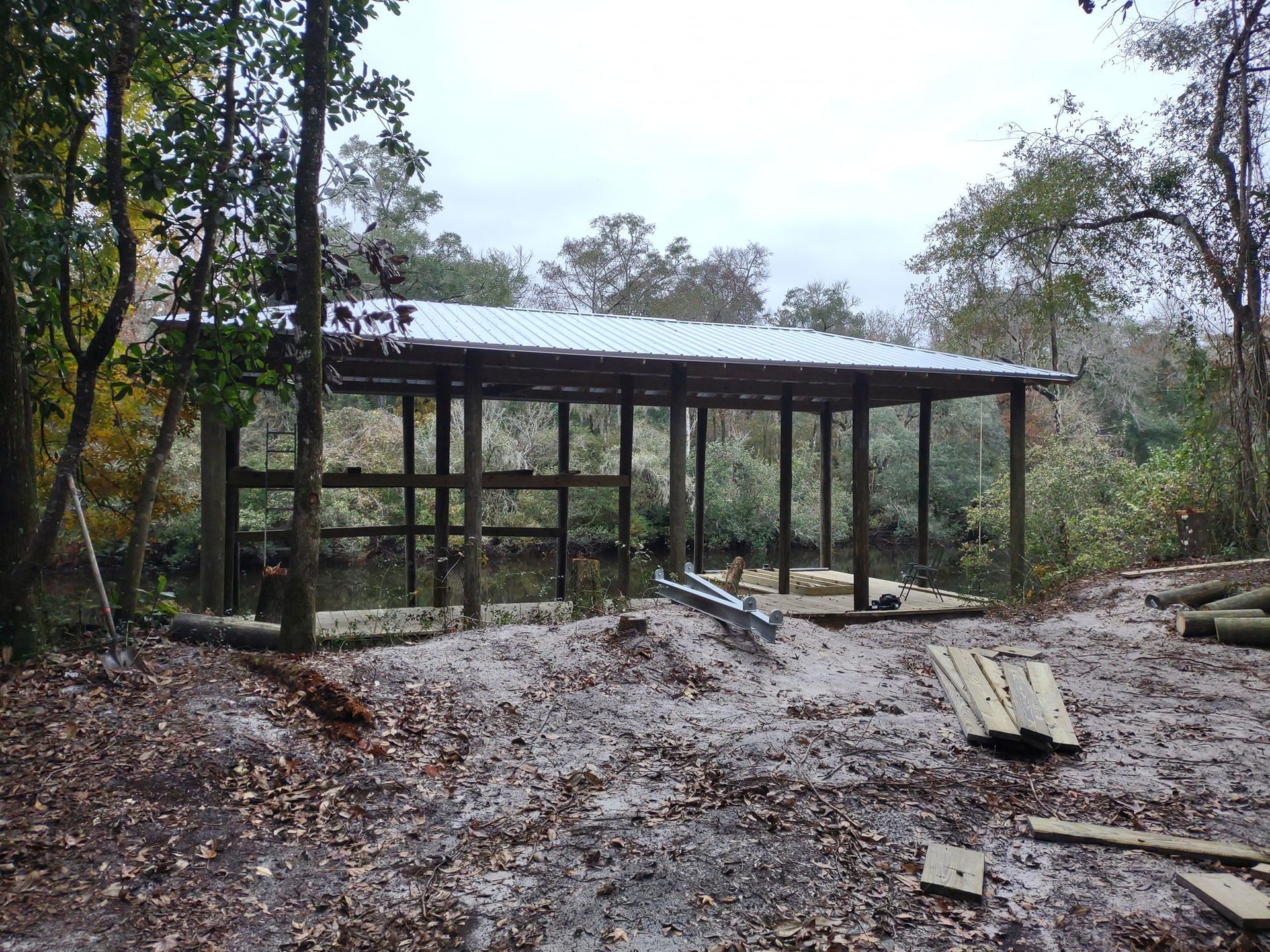 A wooden boat slip structure with a metal roof stands on a forested riverbank surrounded by scattered leaves and lumber.