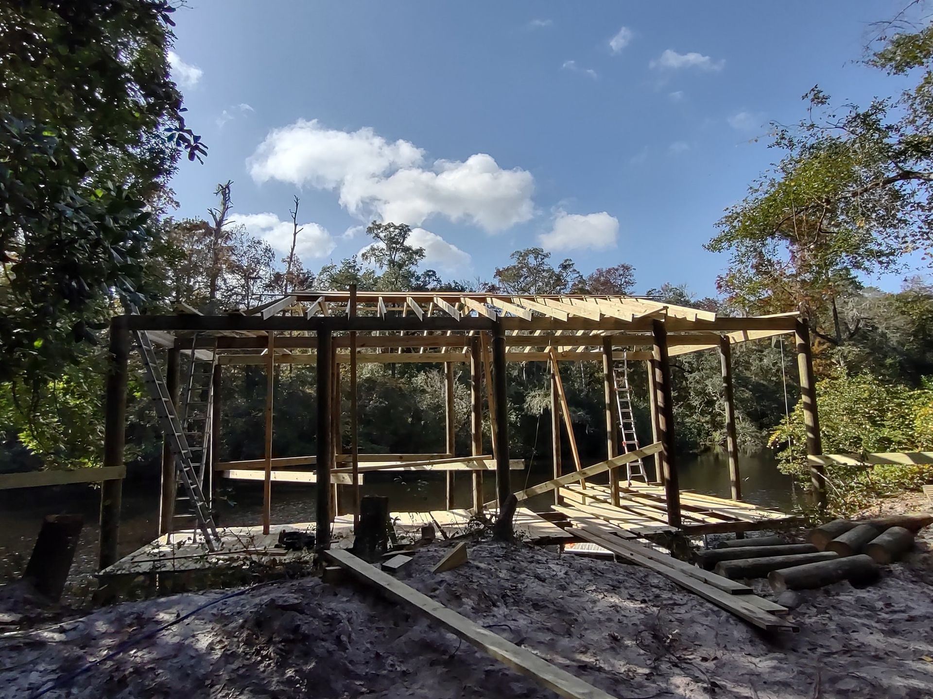 A wooden structure under construction with exposed roof rafters, situated in a wooded area near a small creek.