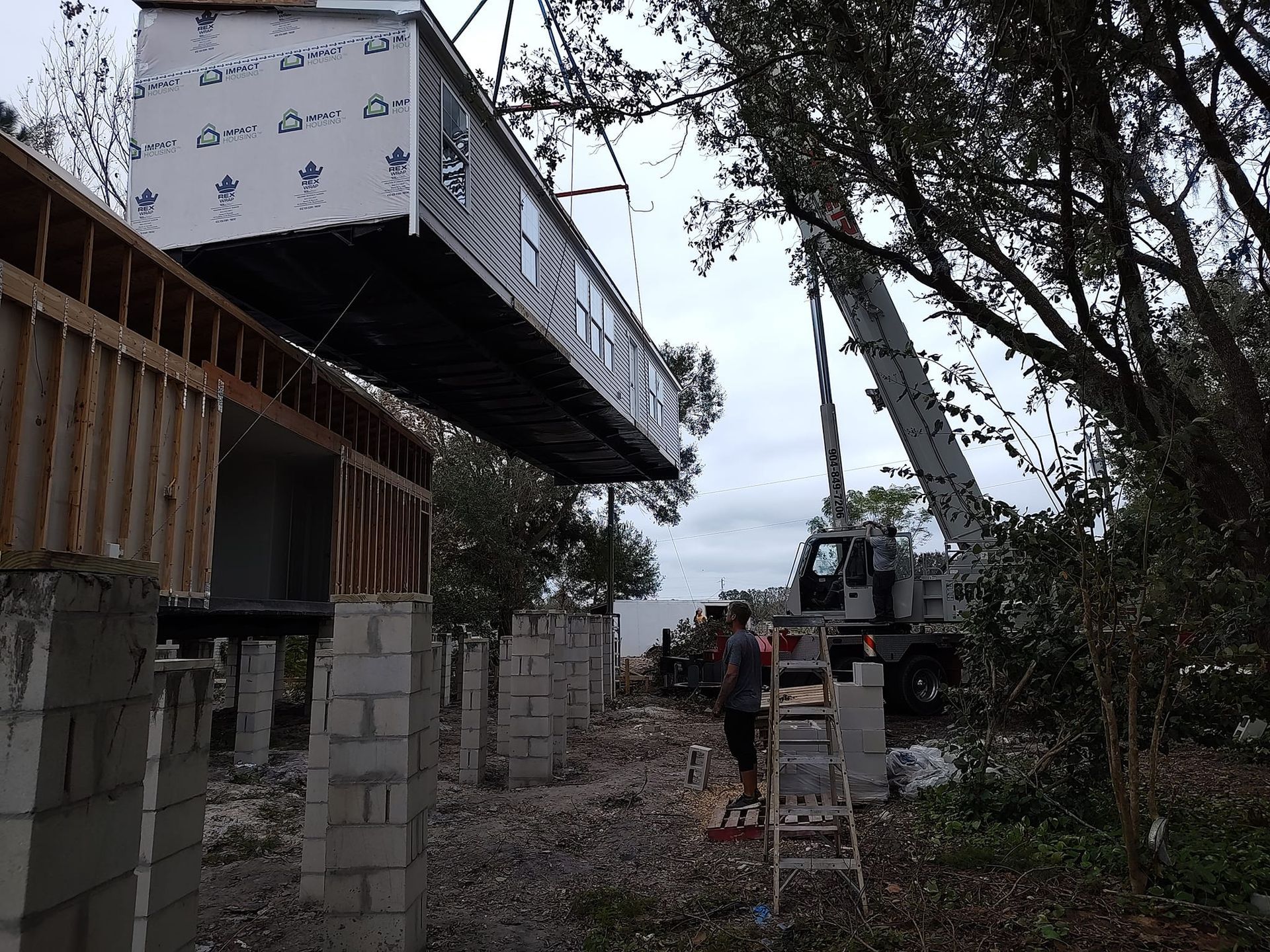 A large crane lifts a section of a modular house onto concrete pillar supports at a construction site.