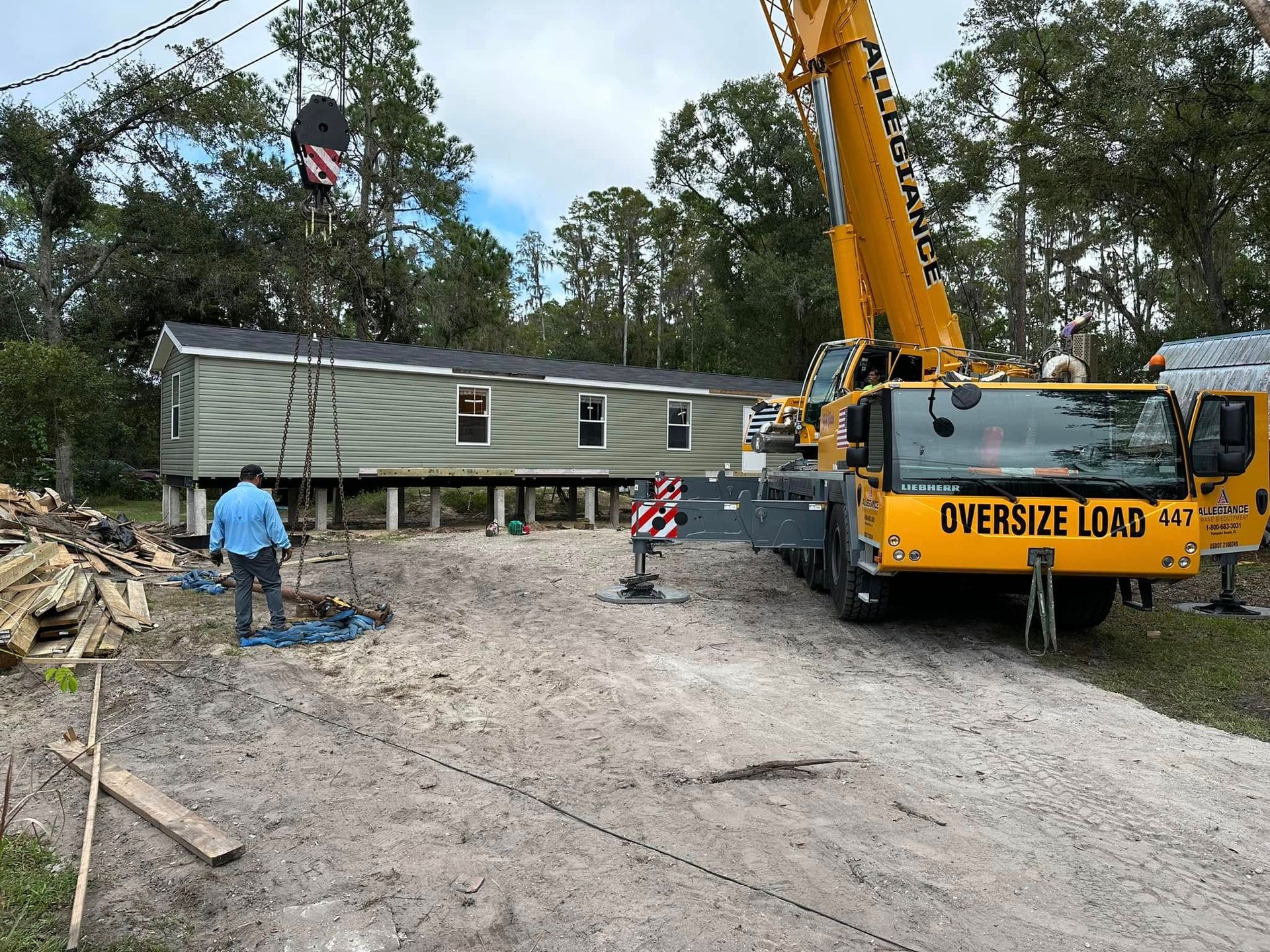 A large yellow crane lifts a mobile home onto a foundation at a construction site surrounded by trees.