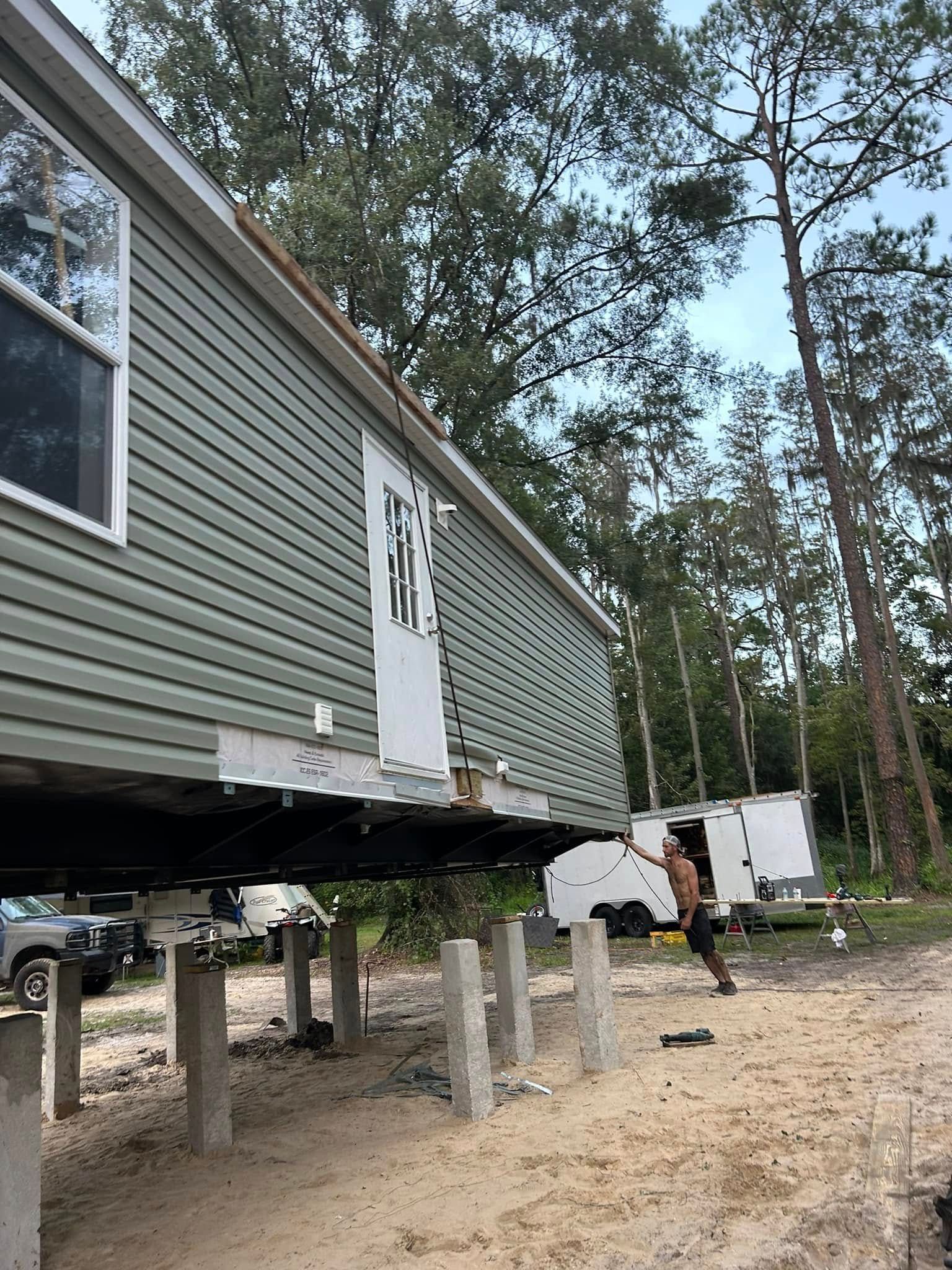A light green mobile home elevated on concrete block piers, with a person standing on the ground beneath the structure.