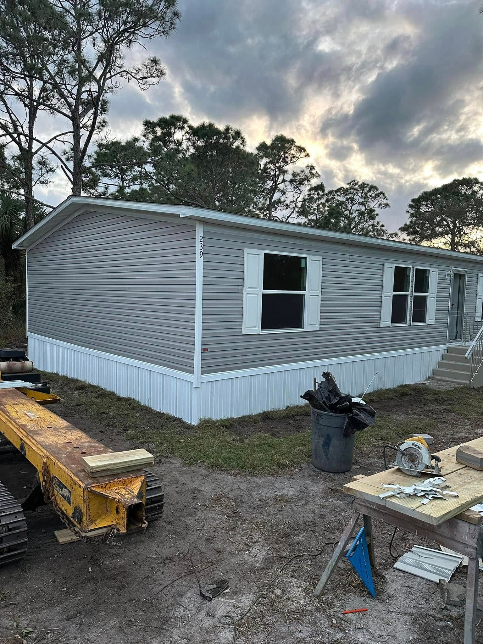A grey manufactured home with a wave-patterned siding, white shutters, and a construction site in the foreground.