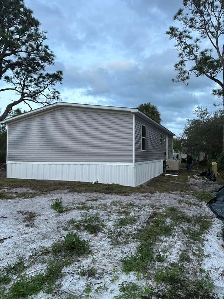 A grey-sided mobile home with white skirting sits on a sandy, partially cleared lot under a cloudy sky.