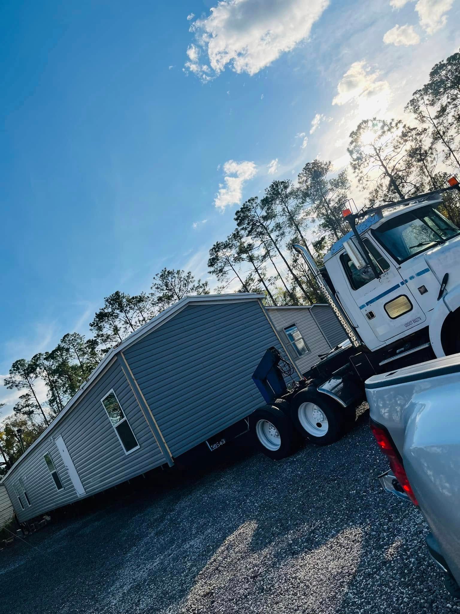 A large, gray manufactured home being towed by a white semi-truck on a gravel lot under a bright, sunny blue sky.
