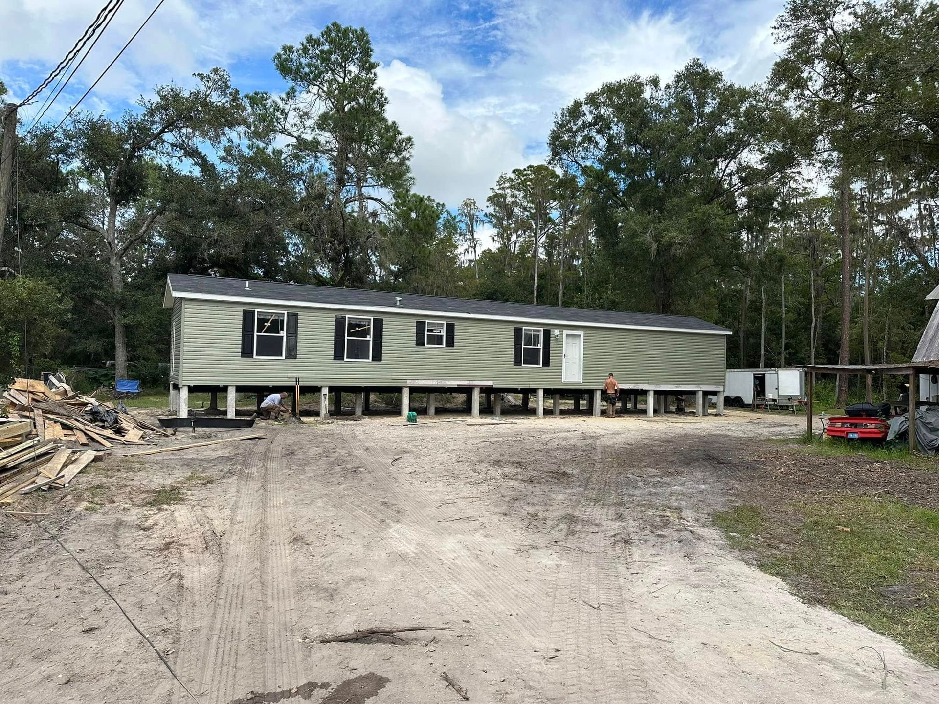 A light green manufactured home on a foundation of white pillars in a wooded, dirt-lot setting.