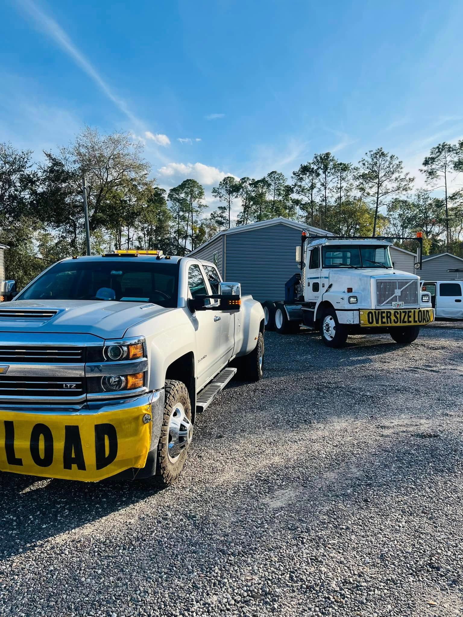 A silver pickup truck and a white transport truck carrying a large shed parked on a gravel lot under a clear blue sky.