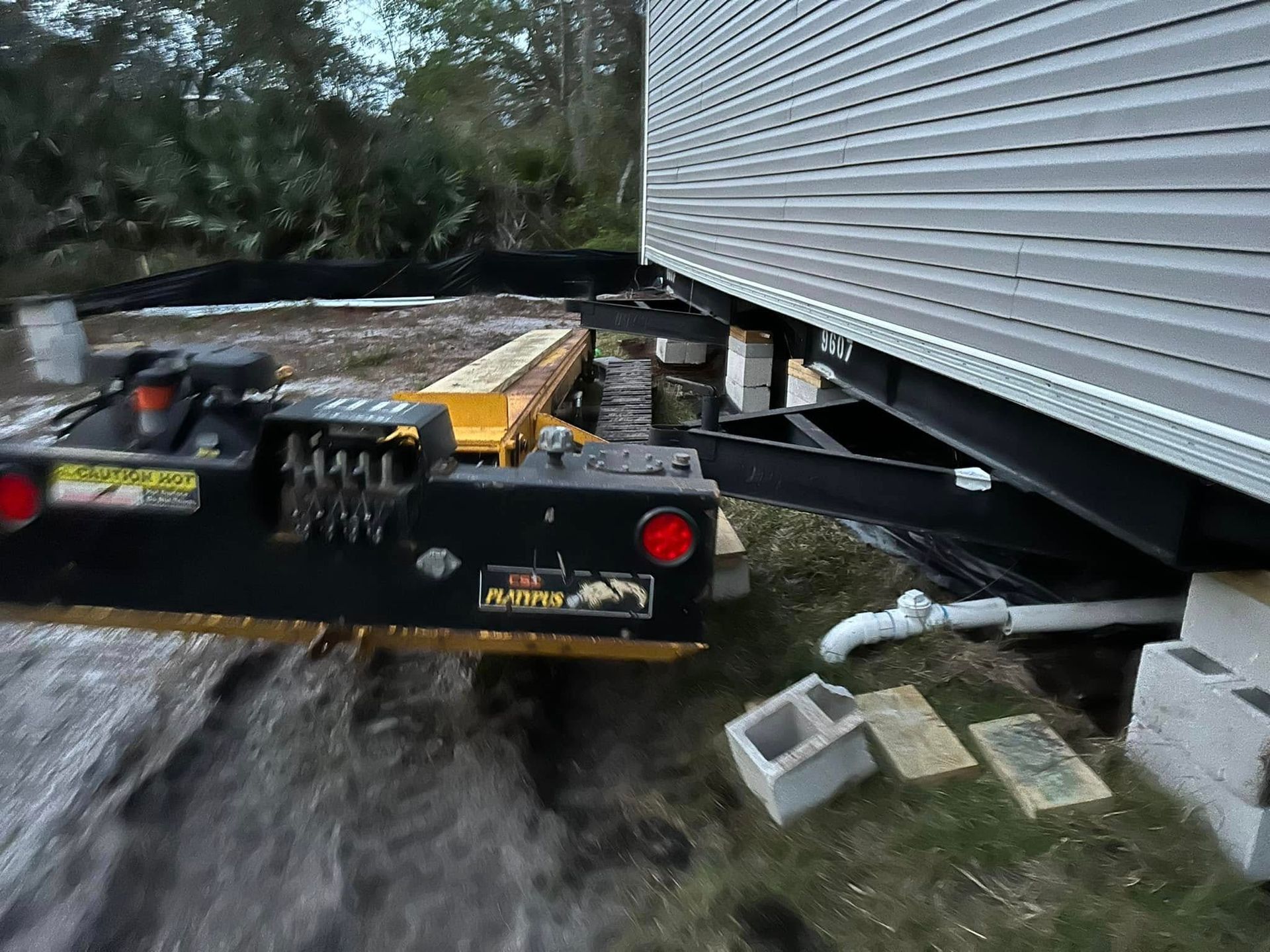 A yellow and black industrial house-moving machine parked next to a mobile home supported by cinder blocks on dirt ground.