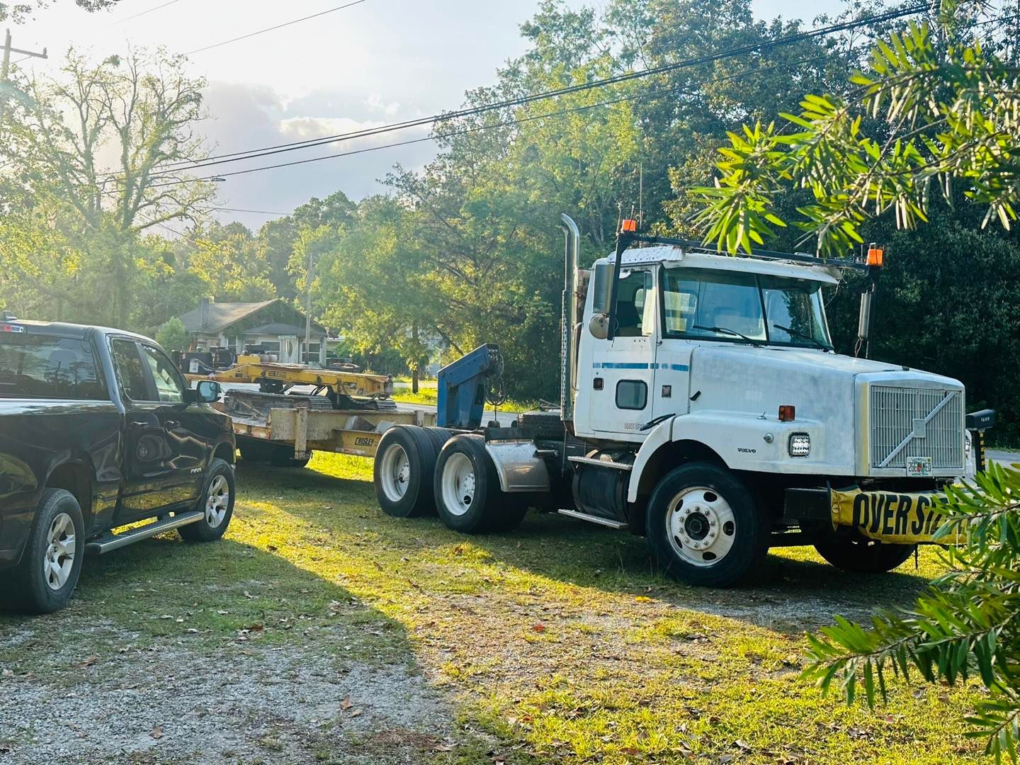 A white semi-truck with an oversized load banner parked next to a black pickup truck on a gravel lot near trees.