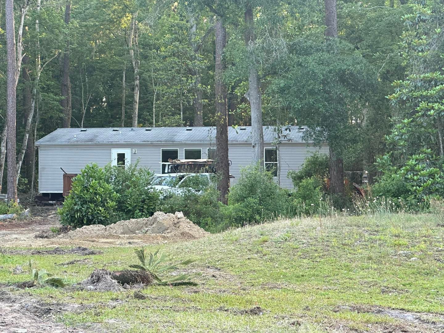 A light-colored mobile home sits in a clearing surrounded by woods, with a white vehicle partially visible near the center.
