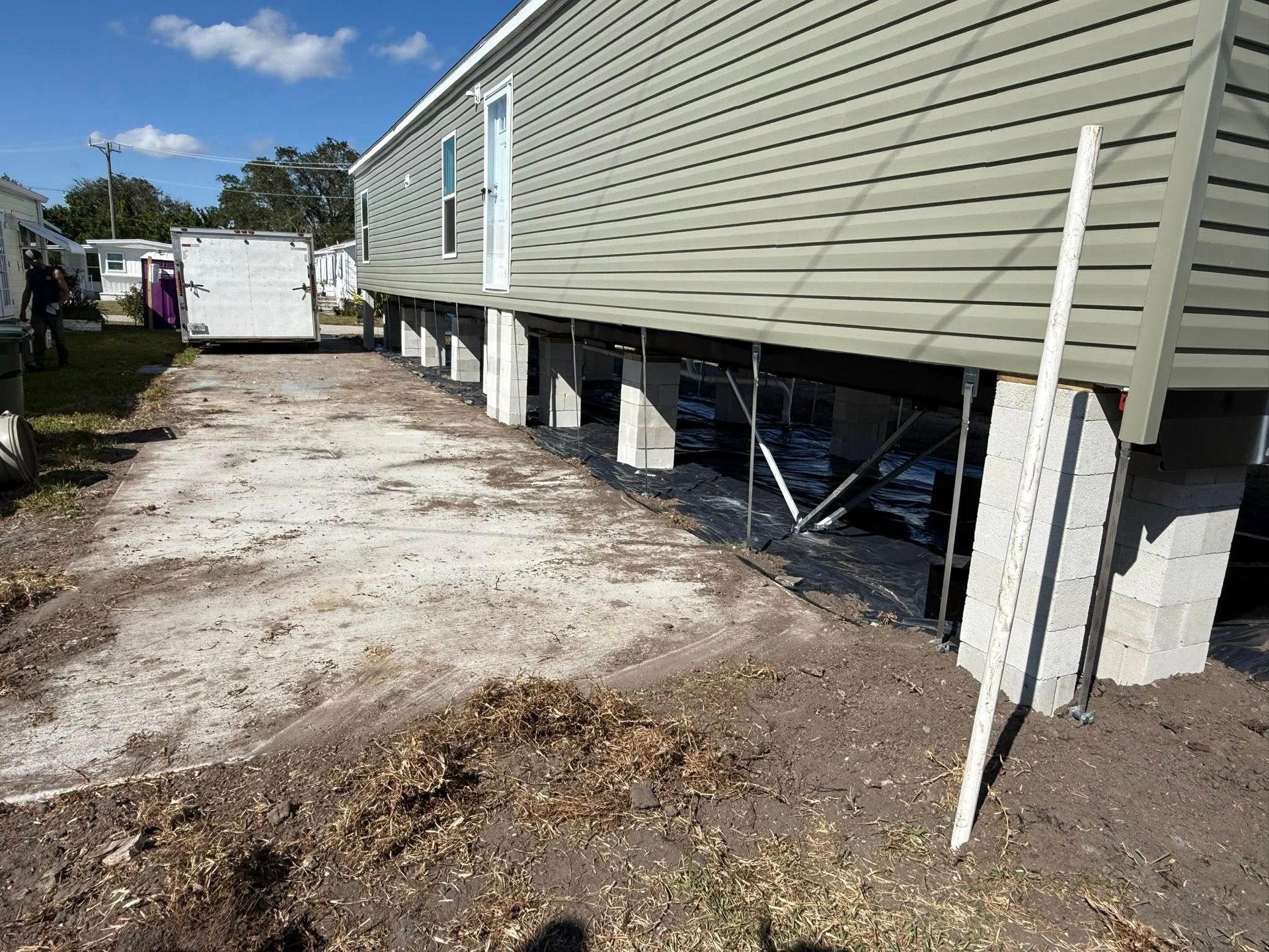 A manufactured home sits on cinder block piers above dirt ground, with a white trailer parked in the background.