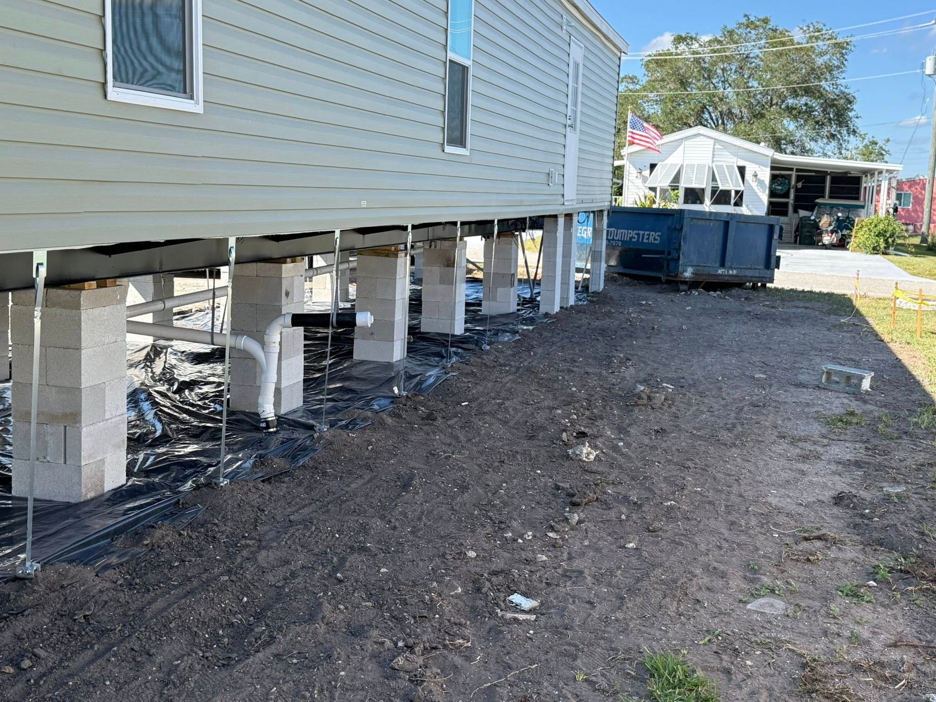 A manufactured home raised on concrete block piers above a ground-level moisture barrier, with a shed in the background.
