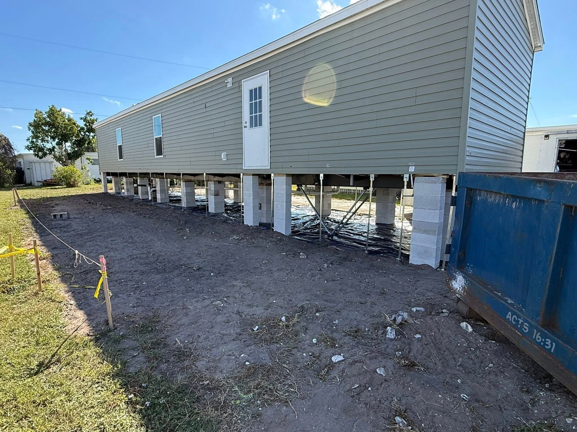 A manufactured home on concrete block piers with green siding, situated on a dirt lot next to a large blue dumpster.