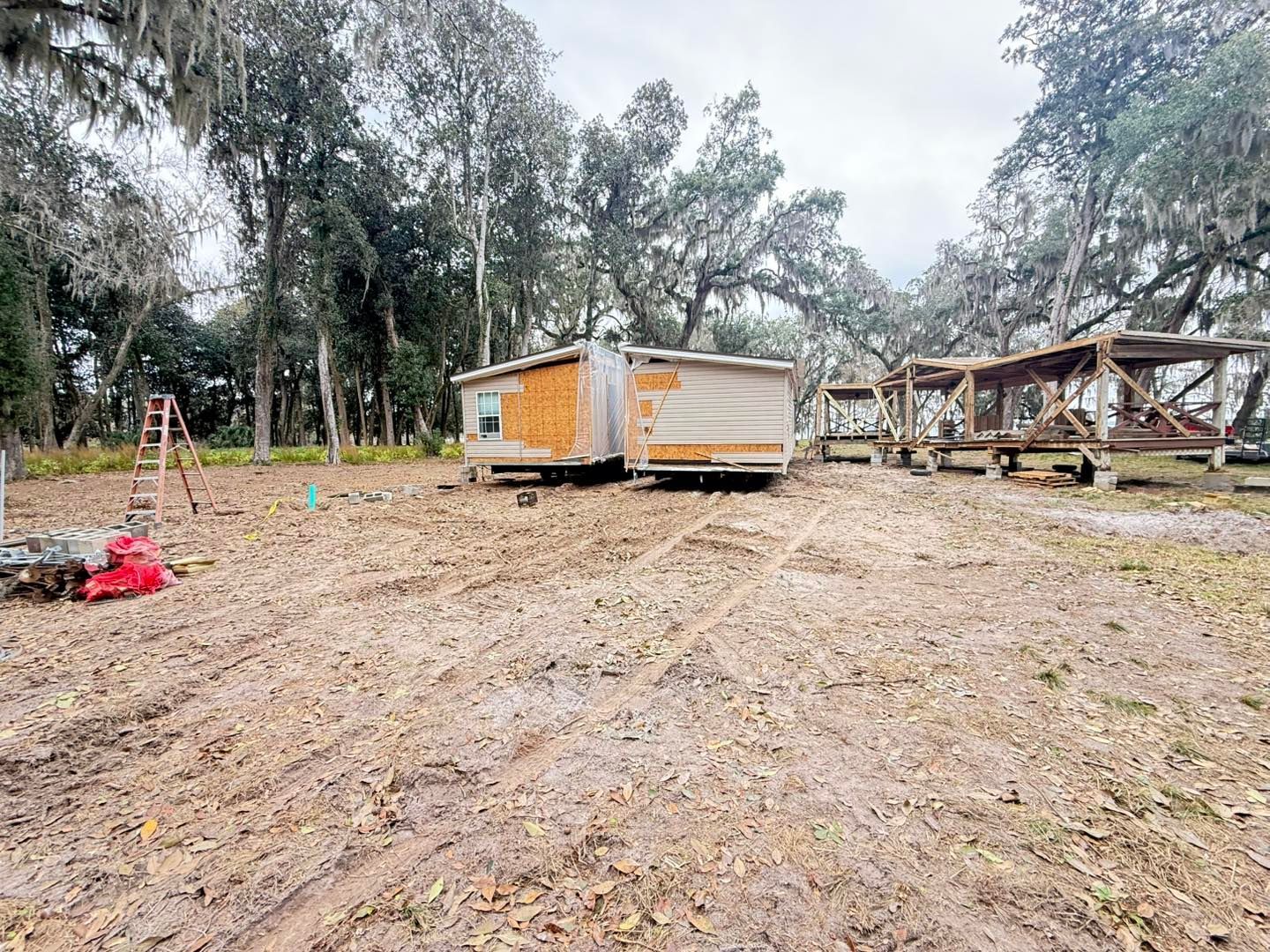 A mobile home under renovation sits in a wooded yard near an open timber-frame structure.