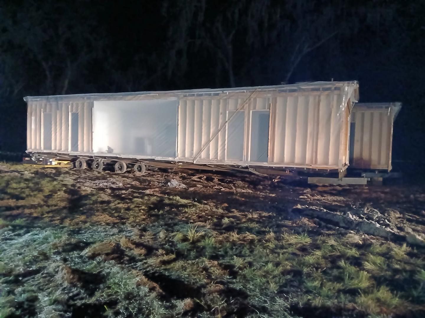 A modular home structure under construction, illuminated at night on a muddy lot.