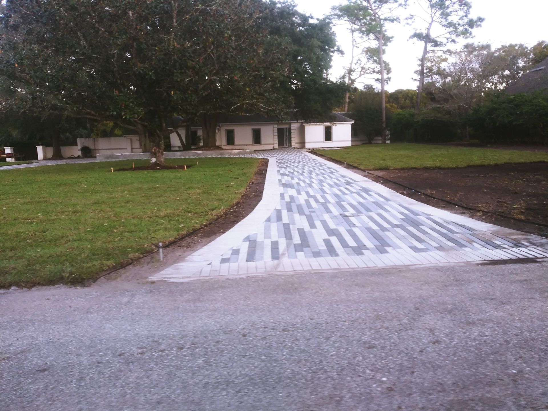 A newly installed, multi-toned gray paved walkway leading toward a white house with a large tree on a grassy lawn.