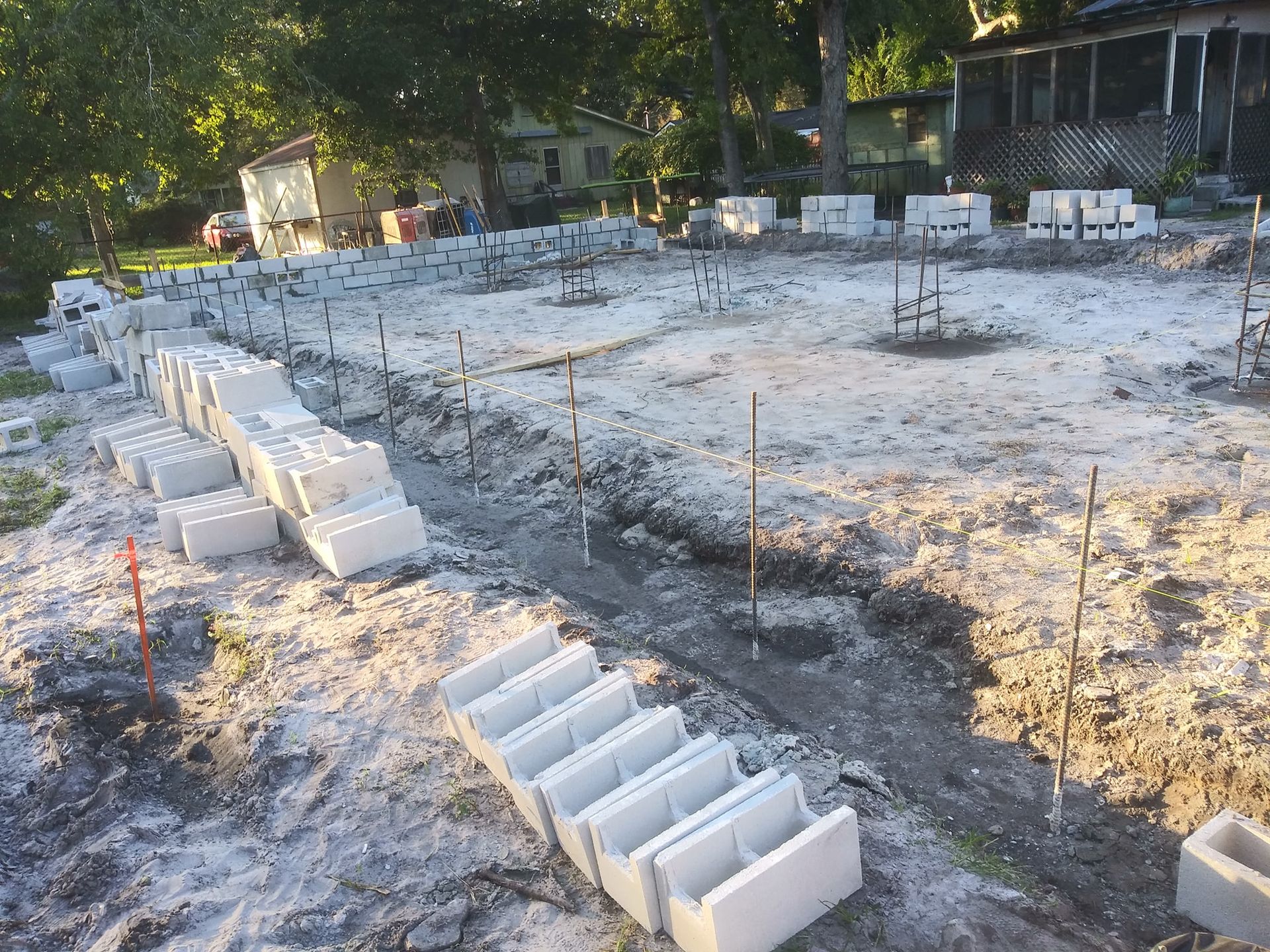 A residential construction site showing a concrete foundation footing, steel rebar, and stacked cinder blocks at dusk.