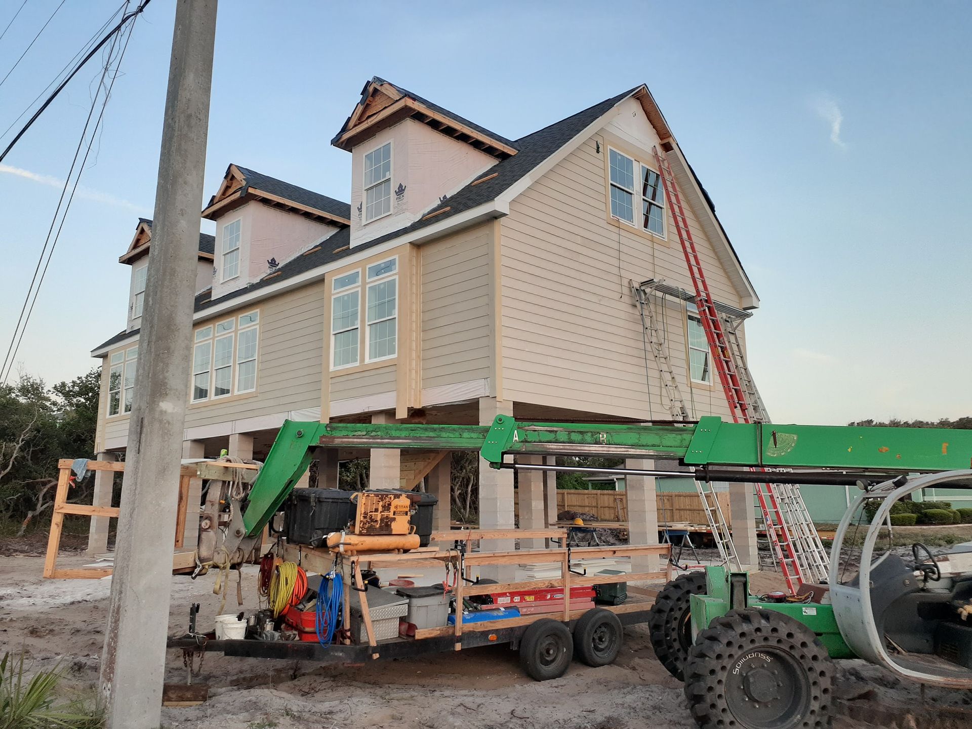 A light-colored house elevated on stilts under construction with an orange ladder, a green lift, and a work trailer below.