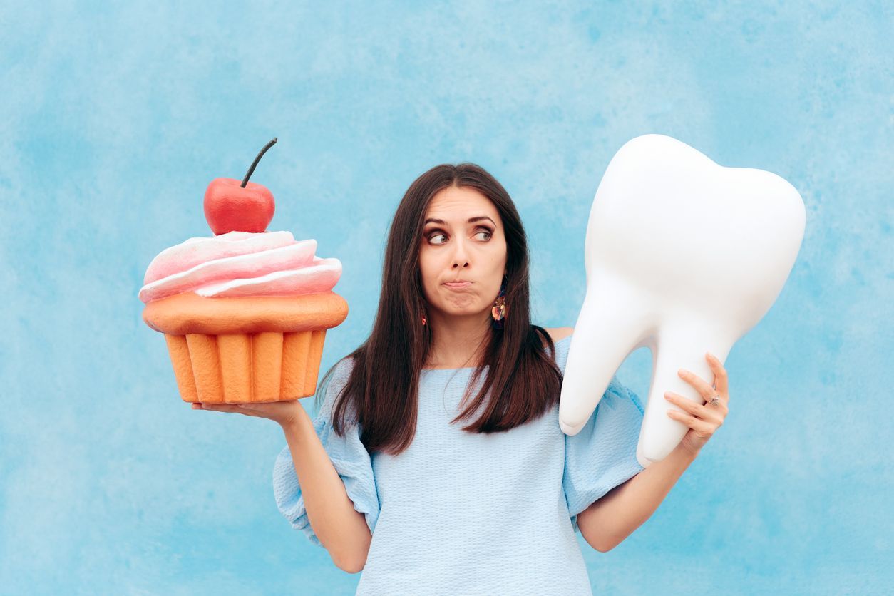 Woman holding giant cupcake and tooth, looking conflicted, against blue backdrop.