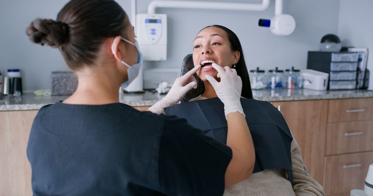 Dentist examining a patient's mouth in a dental office. Both wear masks, patient has mouth open.
