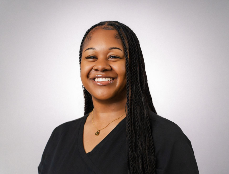 Woman with long braided hair smiles broadly, wearing a black shirt, against a light gray background.