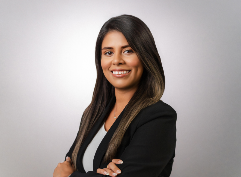 Woman with dark hair, smiling, wearing a black blazer and white top, arms crossed, against a gray background.