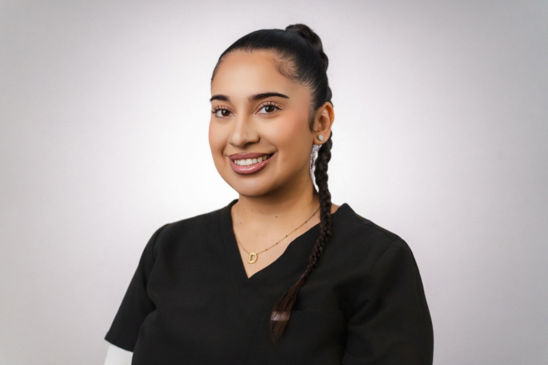 Woman with long dark hair smiles while standing in front of a white brick wall; wearing a dark blue top.