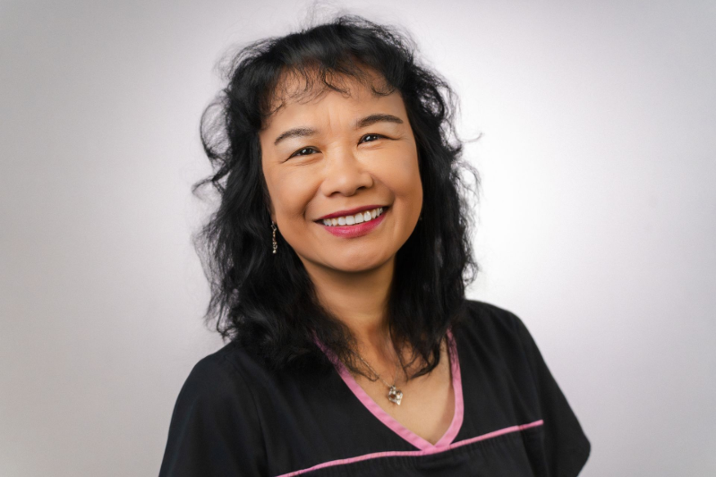 Woman with dark hair smiles, wearing a black shirt with pink trim, against a gray background.