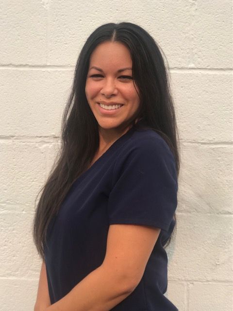 A woman in a blue scrub top is smiling in front of a white wall.