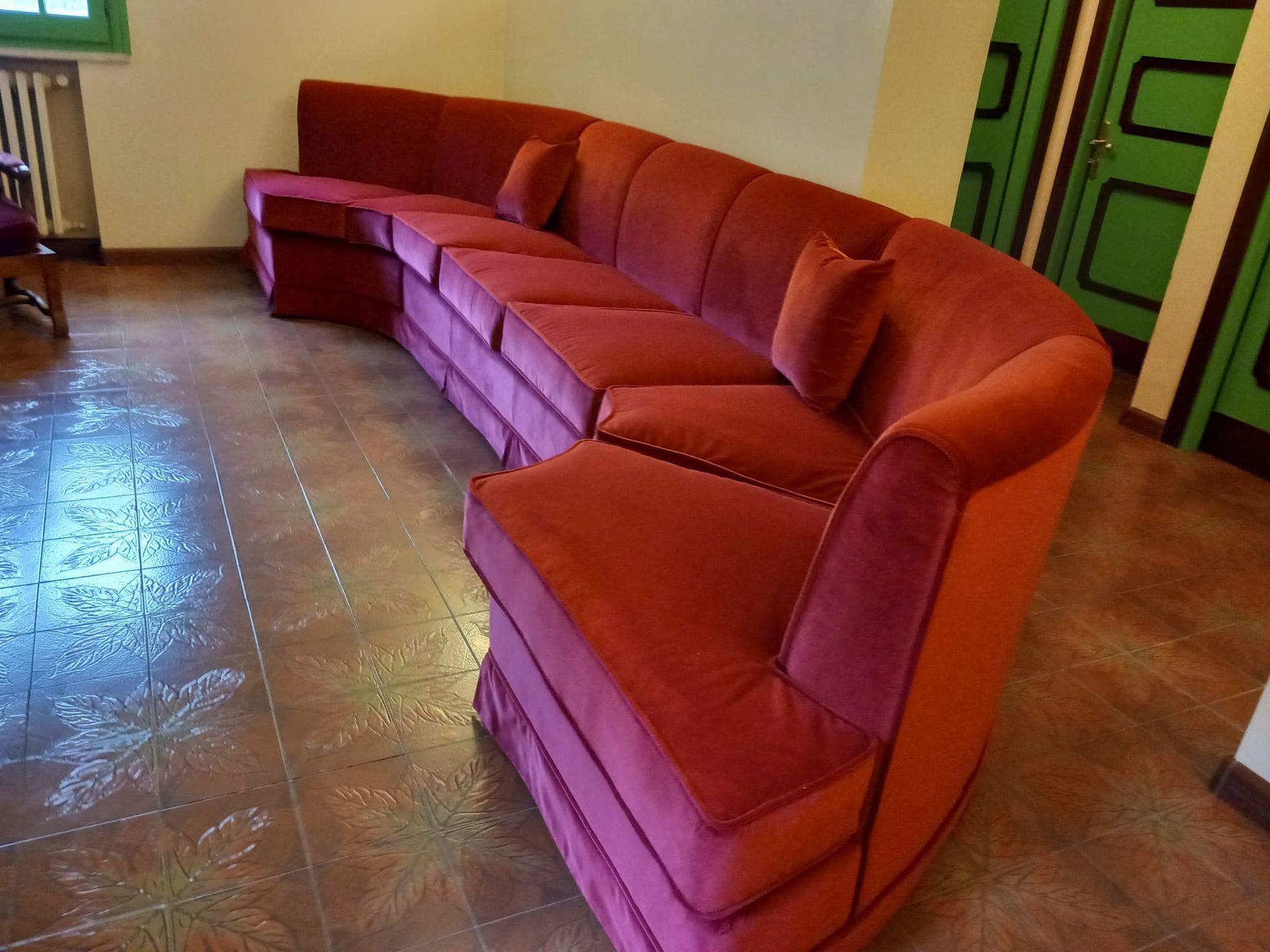 Divano curvo in velluto rosso in una stanza con pavimento in piastrelle a fantasia e porta verde. Sul divano ci sono due cuscini decorativi.
