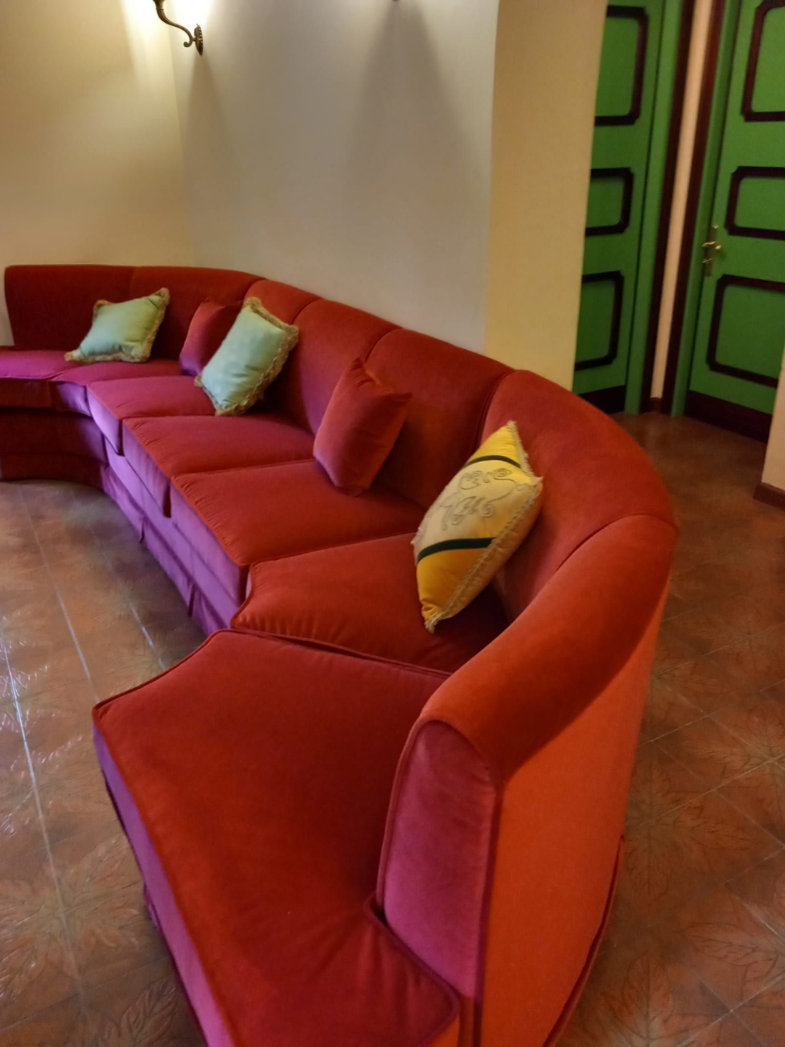 Divano componibile curvo in velluto rosso con cuscini verdi e gialli in una stanza con porte verdi.